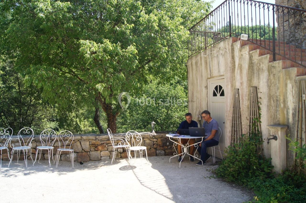 Deux personnes travaillent sur un ordinateur portable à une table en extérieur, près d'un bâtiment en pierre et d'arbres.