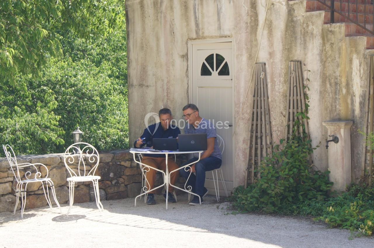 Deux hommes travaillent sur un ordinateur portable à une table en fer forgé, devant une maison en pierre entourée de verdure.