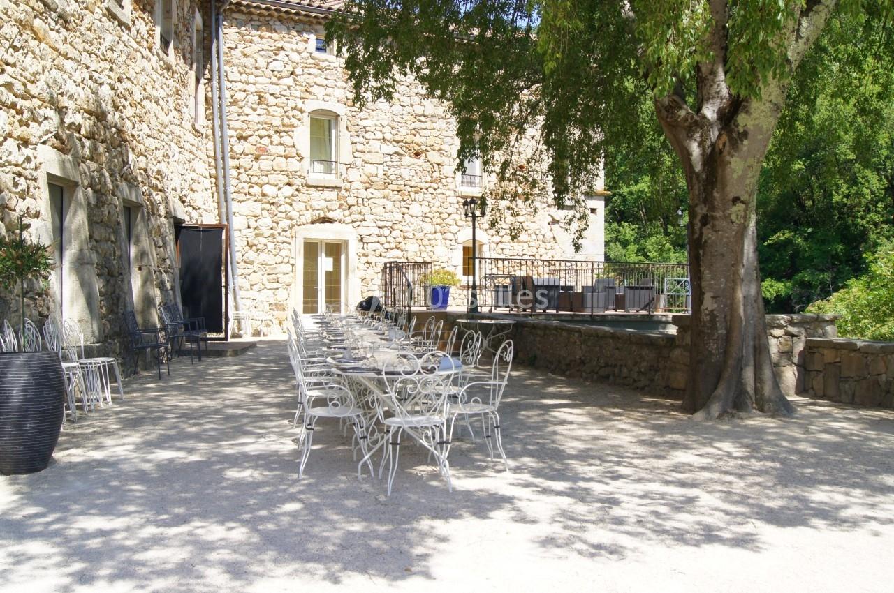Terrasse ensoleillée avec tables et chaises en métal blanc devant un bâtiment en pierre et un grand arbre.