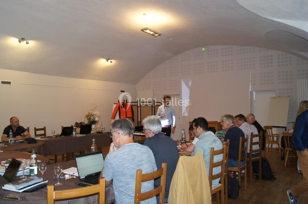 Un groupe de personnes assises autour de tables en réunion dans une salle éclairée avec un présentateur debout.