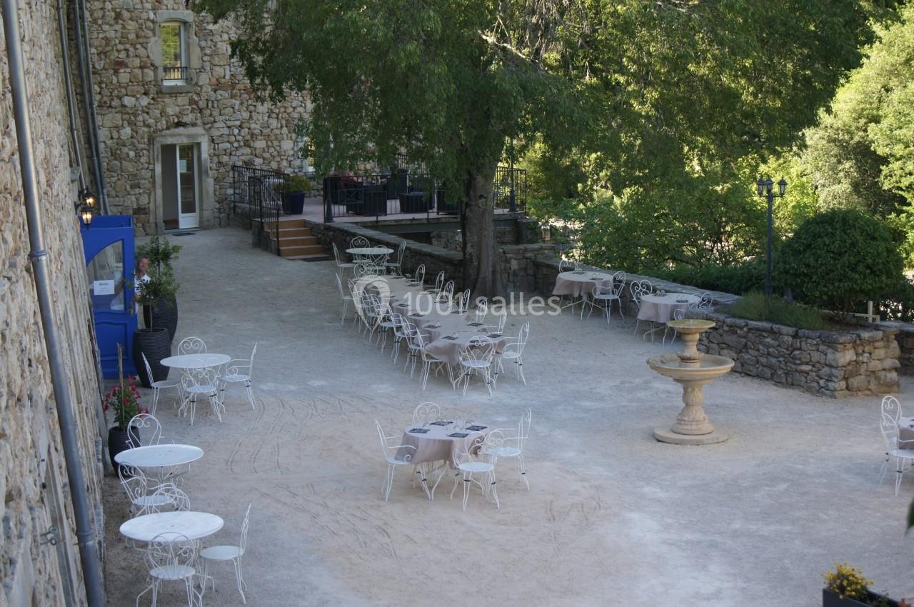 Cour extérieure avec tables et chaises en fer forgé, entourée de verdure et d'un bâtiment en pierre.