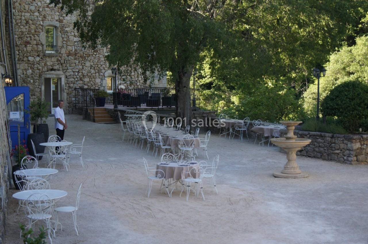 Cour extérieure avec tables et chaises en fer forgé, fontaine centrale et bâtiment en pierre sous des arbres.