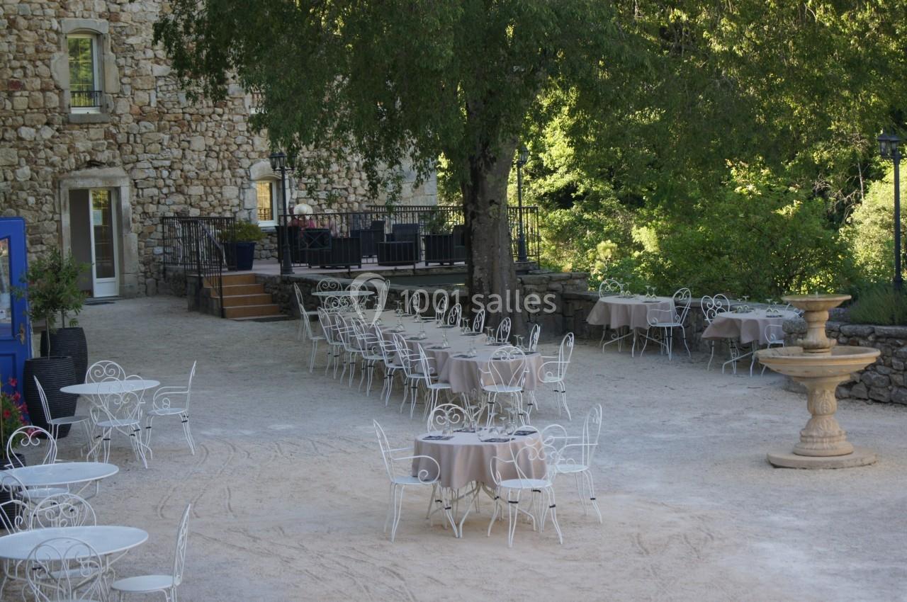 Terrasse extérieure avec tables et chaises en fer forgé, située devant un bâtiment en pierre et entourée de verdure.