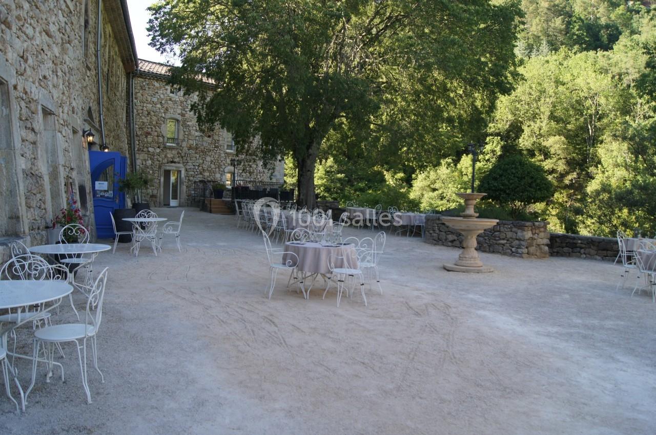 Terrasse en plein air avec tables et chaises en métal blanc, entourée de bâtiments en pierre et de verdure.