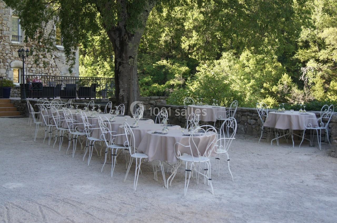 Tables rondes dressées avec nappes beiges et chaises blanches en fer forgé, disposées en extérieur sous un grand arbre.