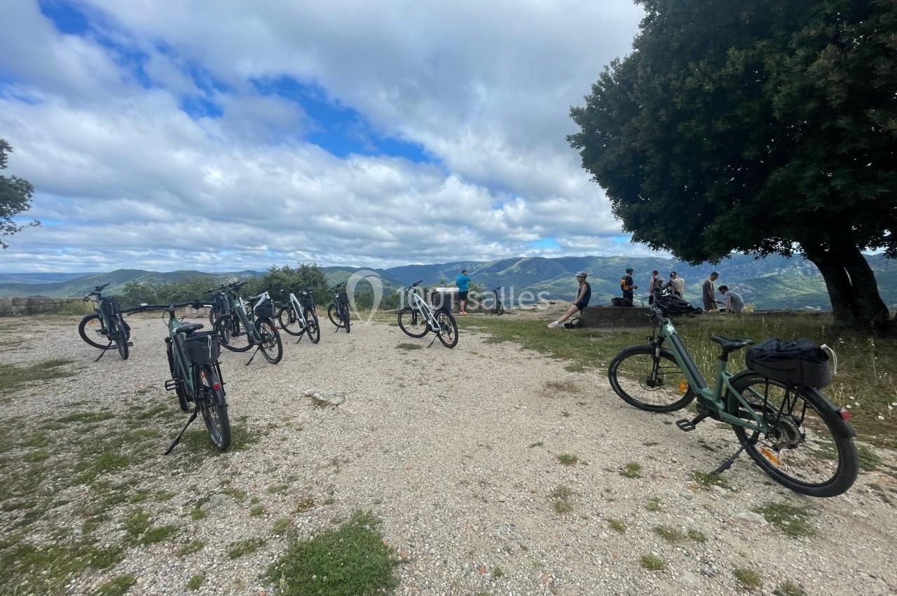 Vélos stationnés sur un chemin de gravier avec des personnes assises sous un arbre, vue sur des collines en arrière-plan.