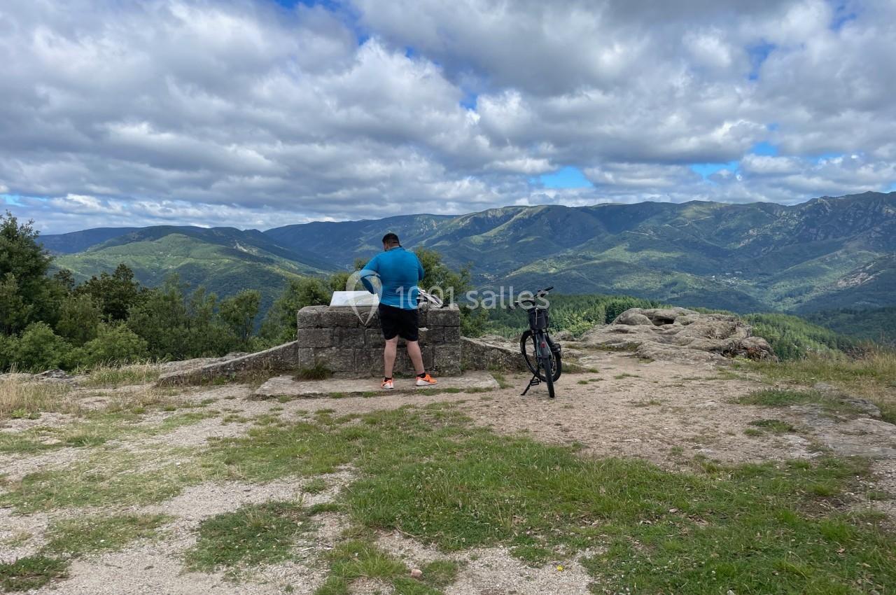Un homme debout près d'une table d'orientation en pierre, avec un vélo posé à côté, face à un paysage montagneux.