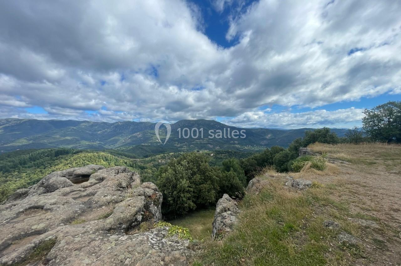 Paysage de montagne avec des rochers au premier plan, une vallée verdoyante et un ciel partiellement nuageux.
