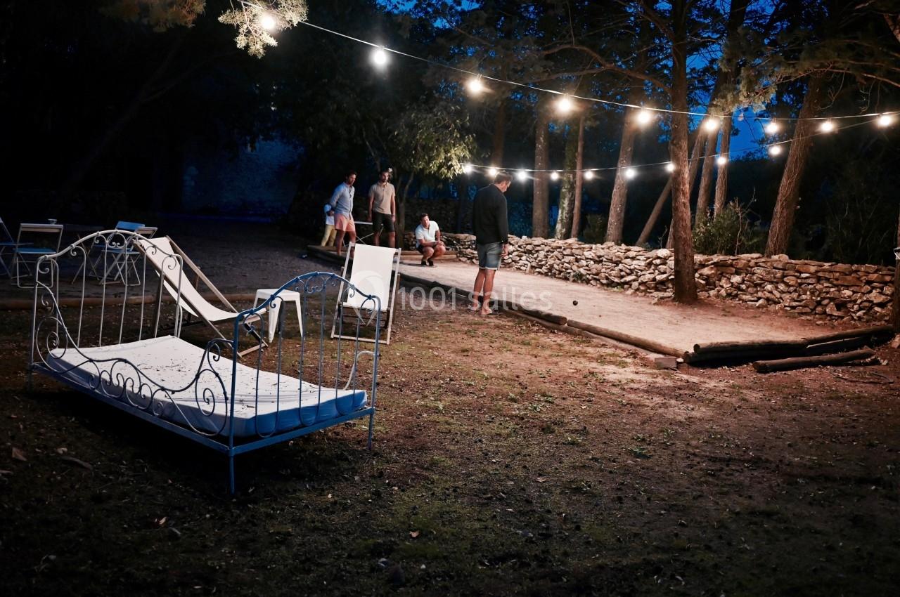 Lit en fer forgé placé en extérieur sous des guirlandes lumineuses, avec des personnes discutant en arrière-plan.