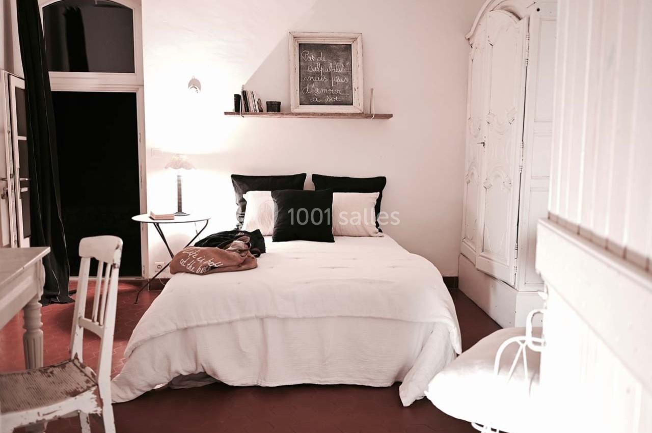 Chambre lumineuse avec lit double, draps blancs, coussins noirs, armoire ancienne et tableau décoratif au mur.