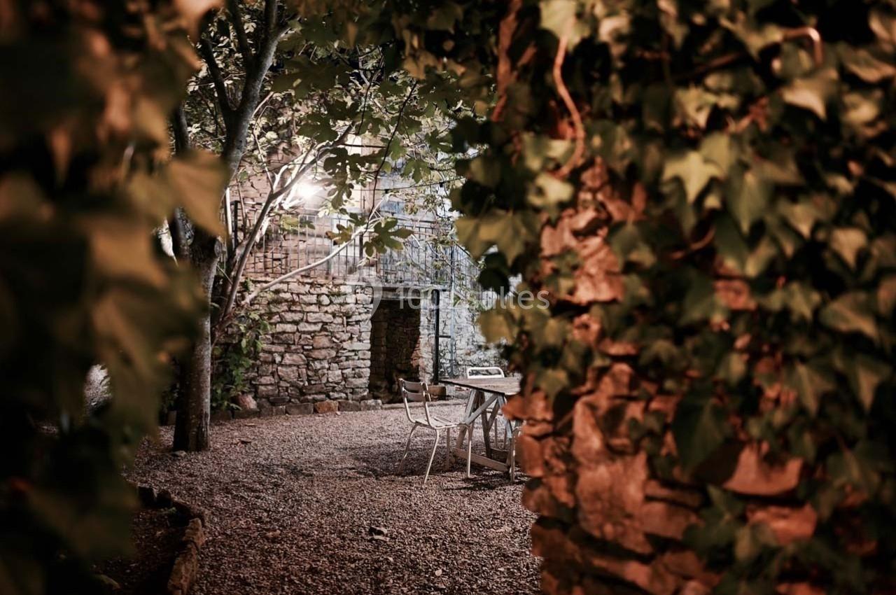 Cour extérieure en soirée, entourée de murs en pierre et de végétation, avec une table et des chaises sur un sol gravillonné.