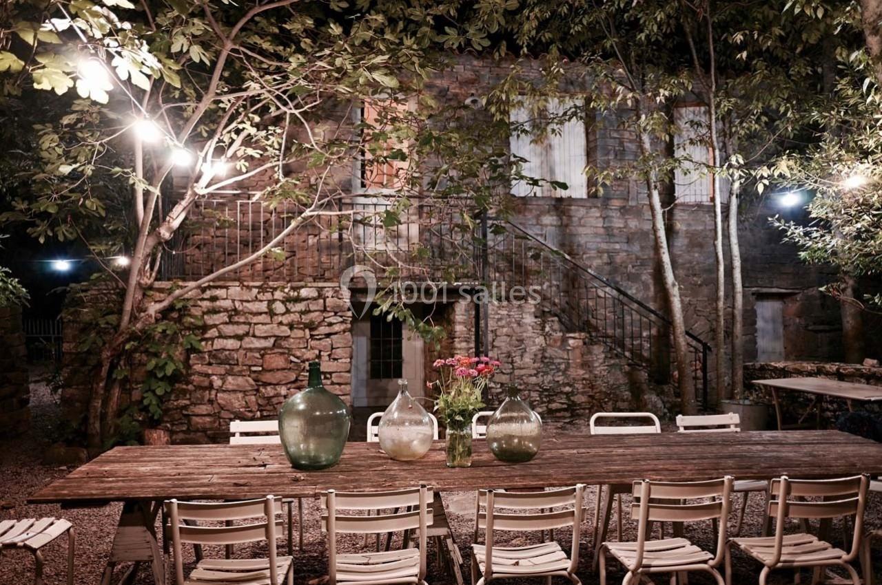 Grande table en bois entourée de chaises blanches dans une cour extérieure éclairée, avec des arbres et un mur en pierre.