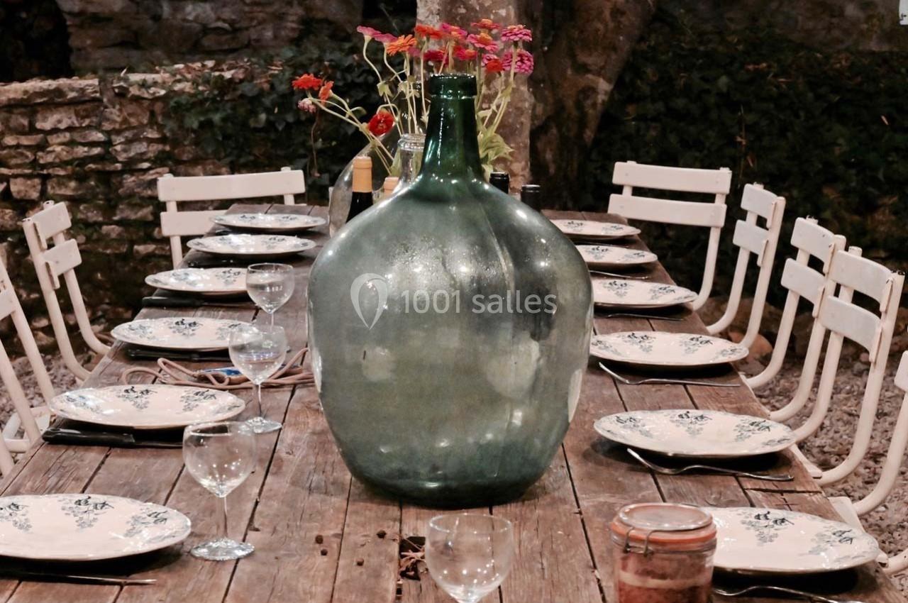 Grande table en bois dressée avec assiettes, verres et une bonbonne en verre vert au centre, entourée de chaises blanches.