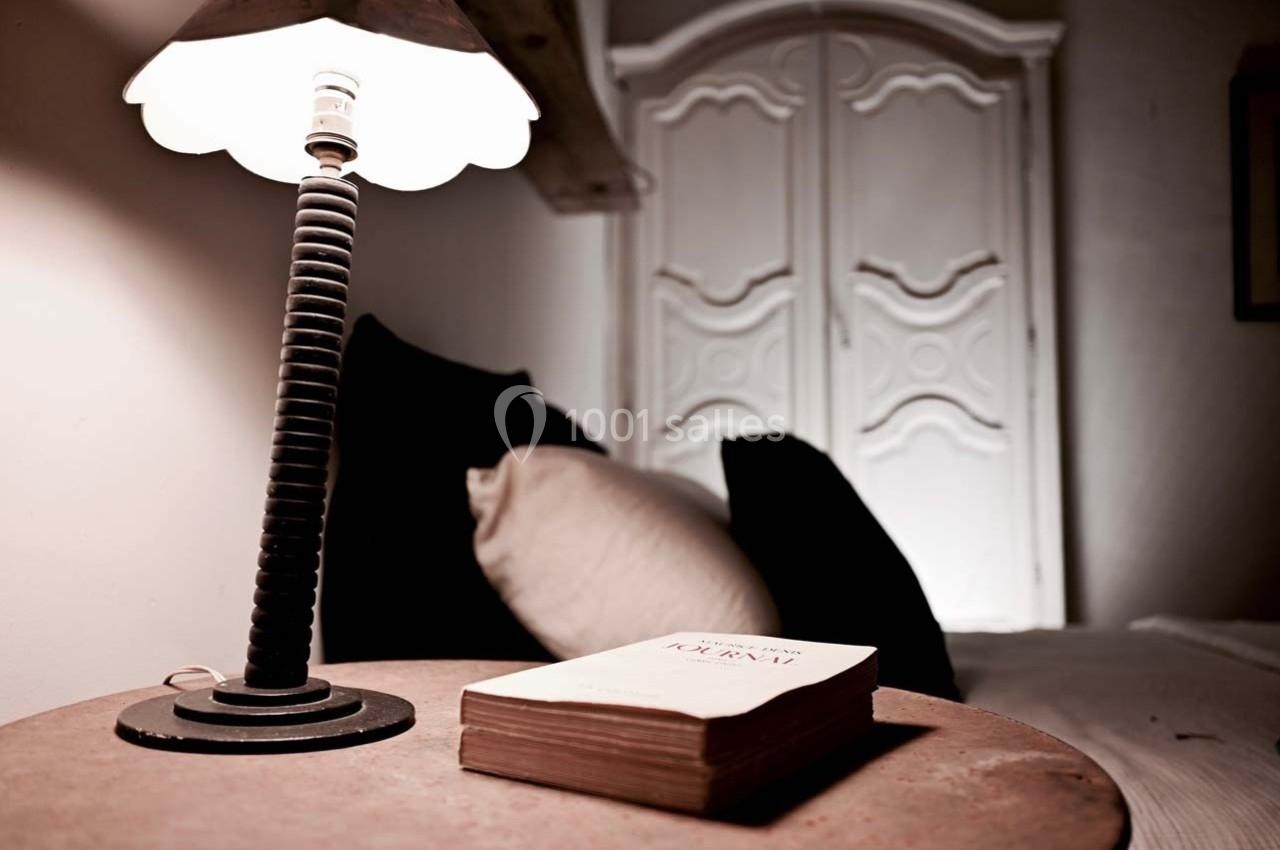 Lampe allumée sur une table en bois avec un livre, devant un lit et une armoire blanche en arrière-plan.