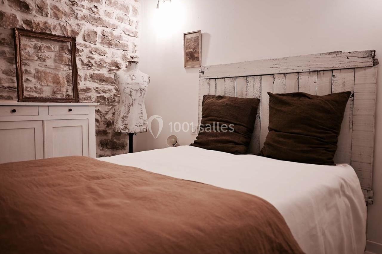 Lit double avec draps blancs et couverture marron dans une chambre au style rustique avec mur en pierre apparente.