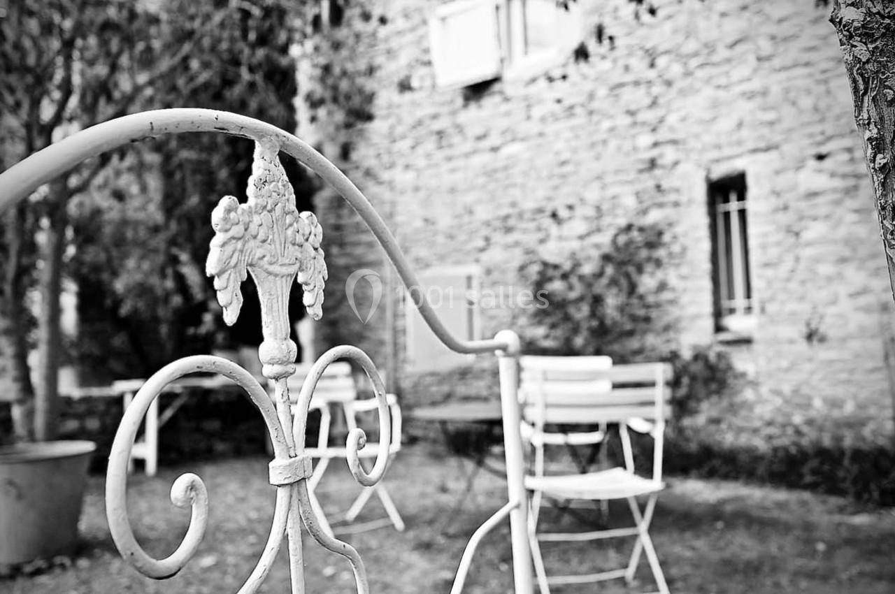 Vue en noir et blanc d'un jardin avec une clôture métallique décorative, des chaises et une façade en pierre.