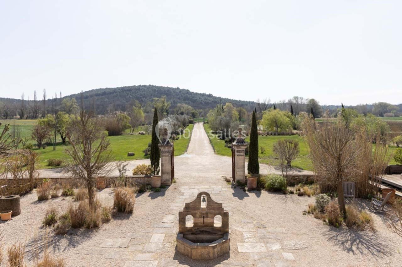 Allée bordée de végétation menant à un paysage vallonné, avec une fontaine en pierre au premier plan.