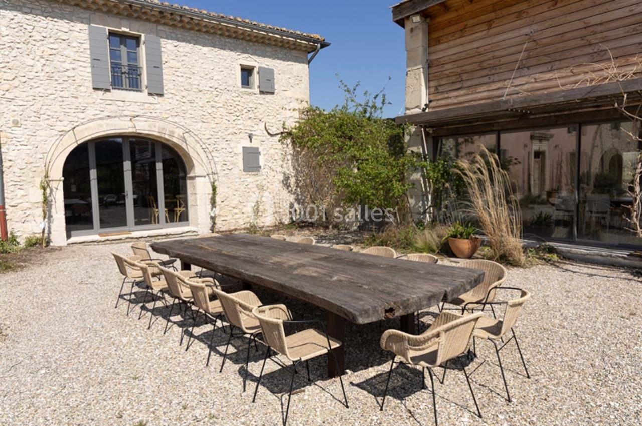 Table en bois massif entourée de chaises en osier dans une cour en gravier, entre deux bâtiments en pierre et bois.