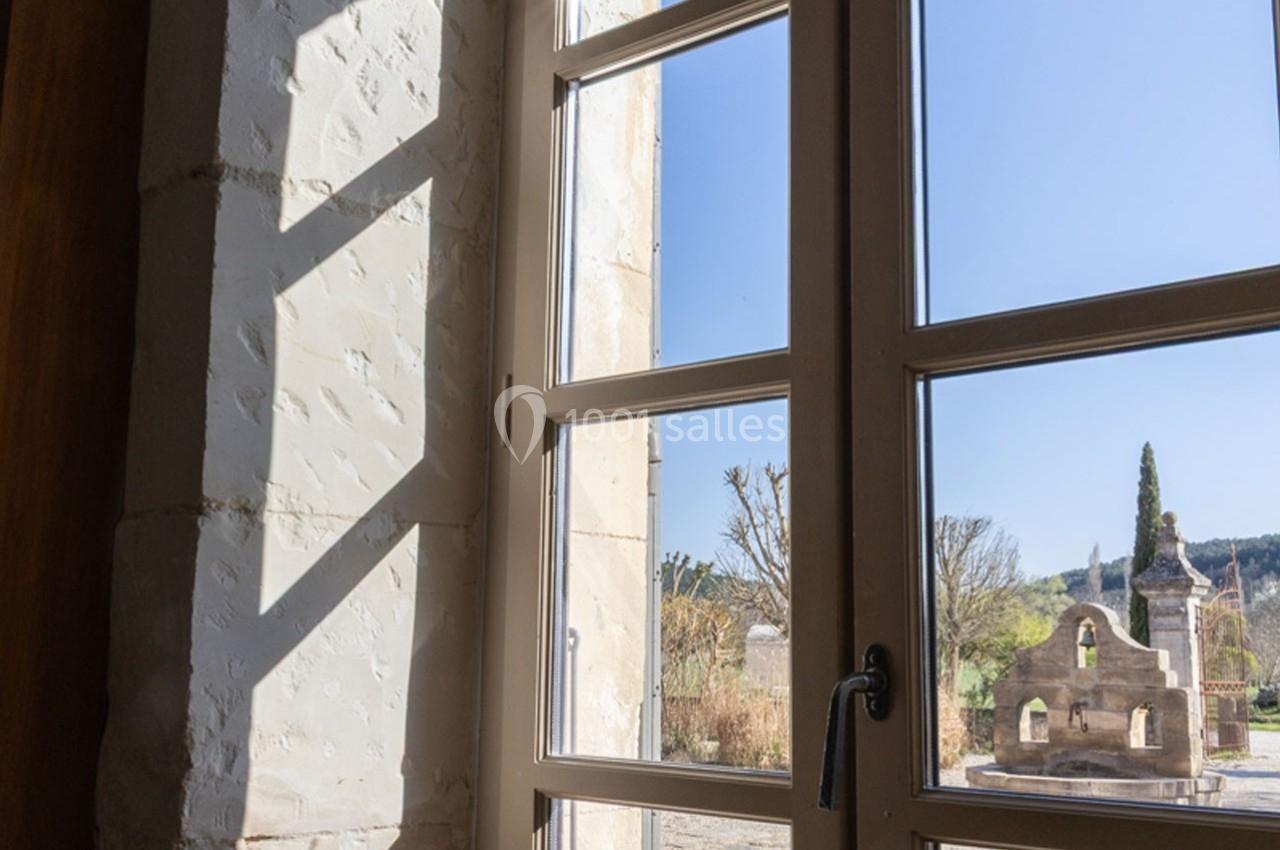 Vue depuis une fenêtre en bois donnant sur un jardin avec une petite structure en pierre et un ciel dégagé.