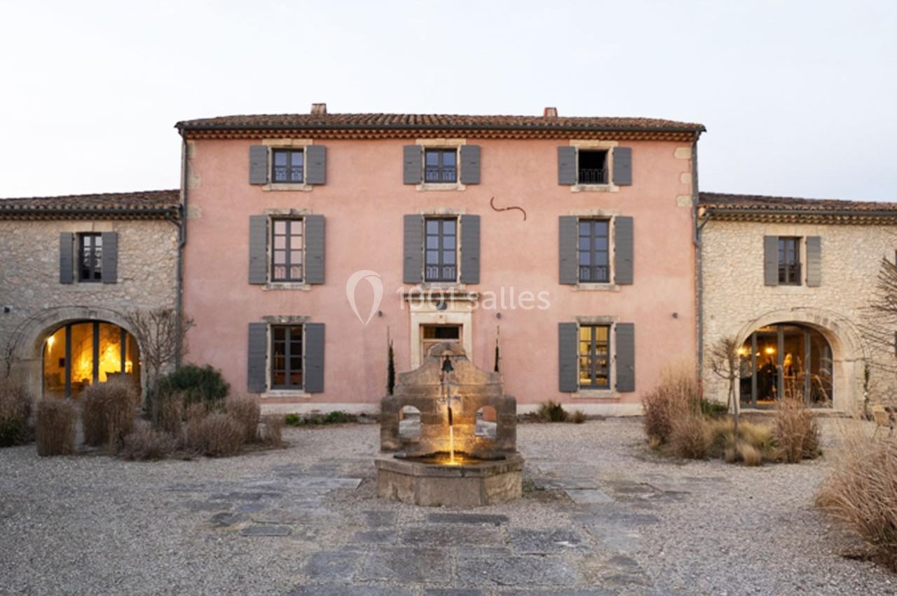 Façade d'une maison en pierre rose avec volets gris, une fontaine au premier plan et un jardin gravillonné.
