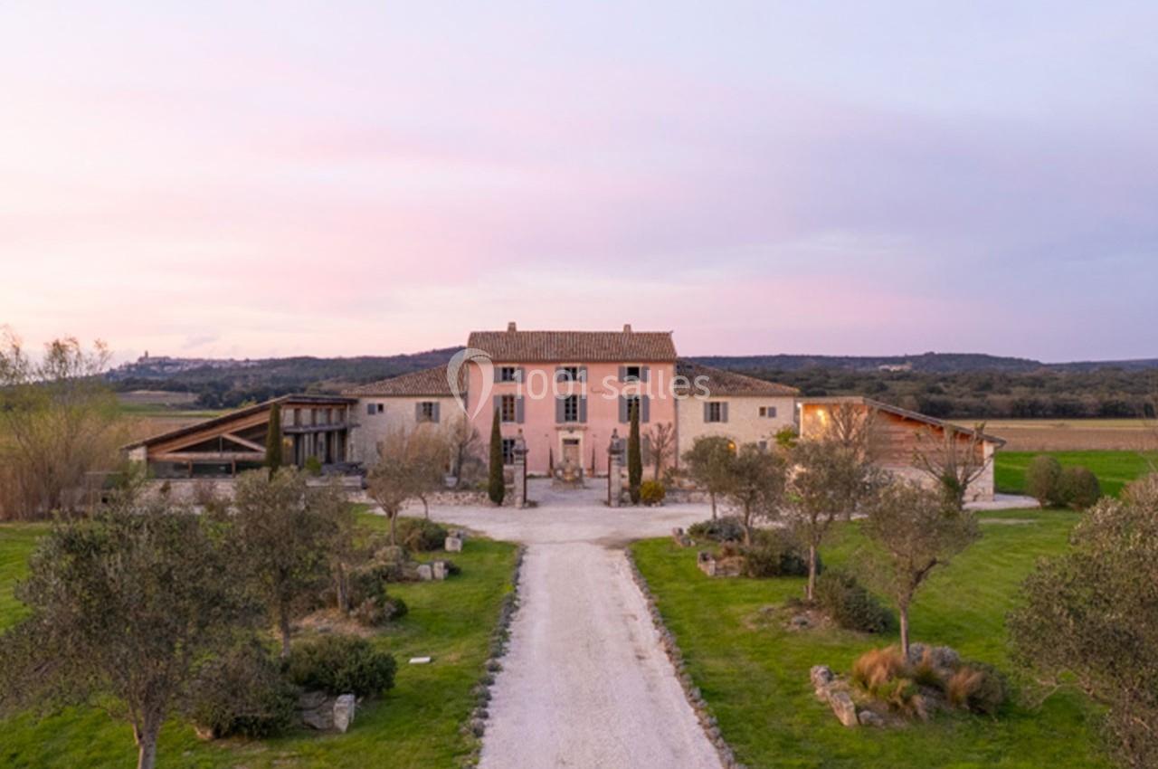 Allée bordée d'oliviers menant à une grande maison en pierre au crépuscule, entourée de champs et de collines.