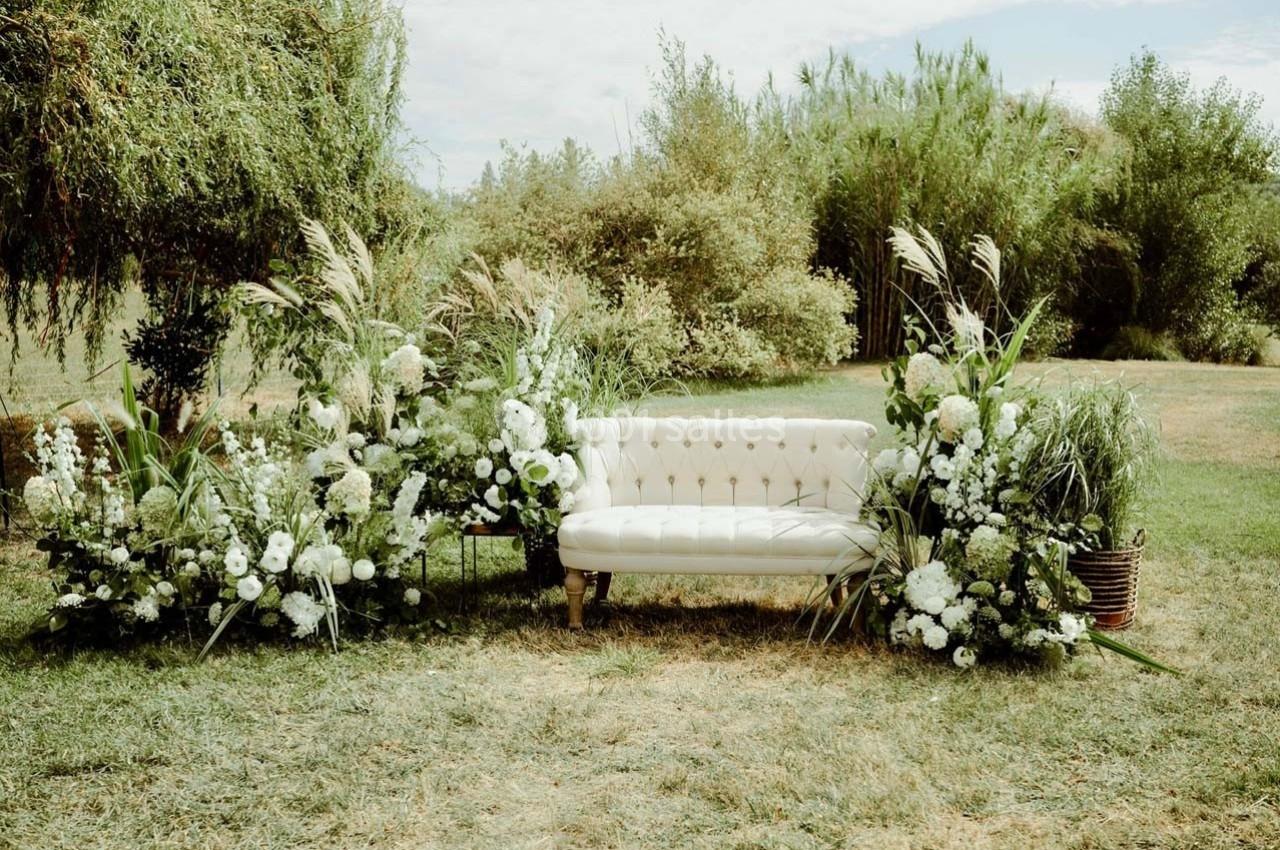 Canapé blanc entouré de compositions florales blanches et vertes dans un jardin verdoyant.
