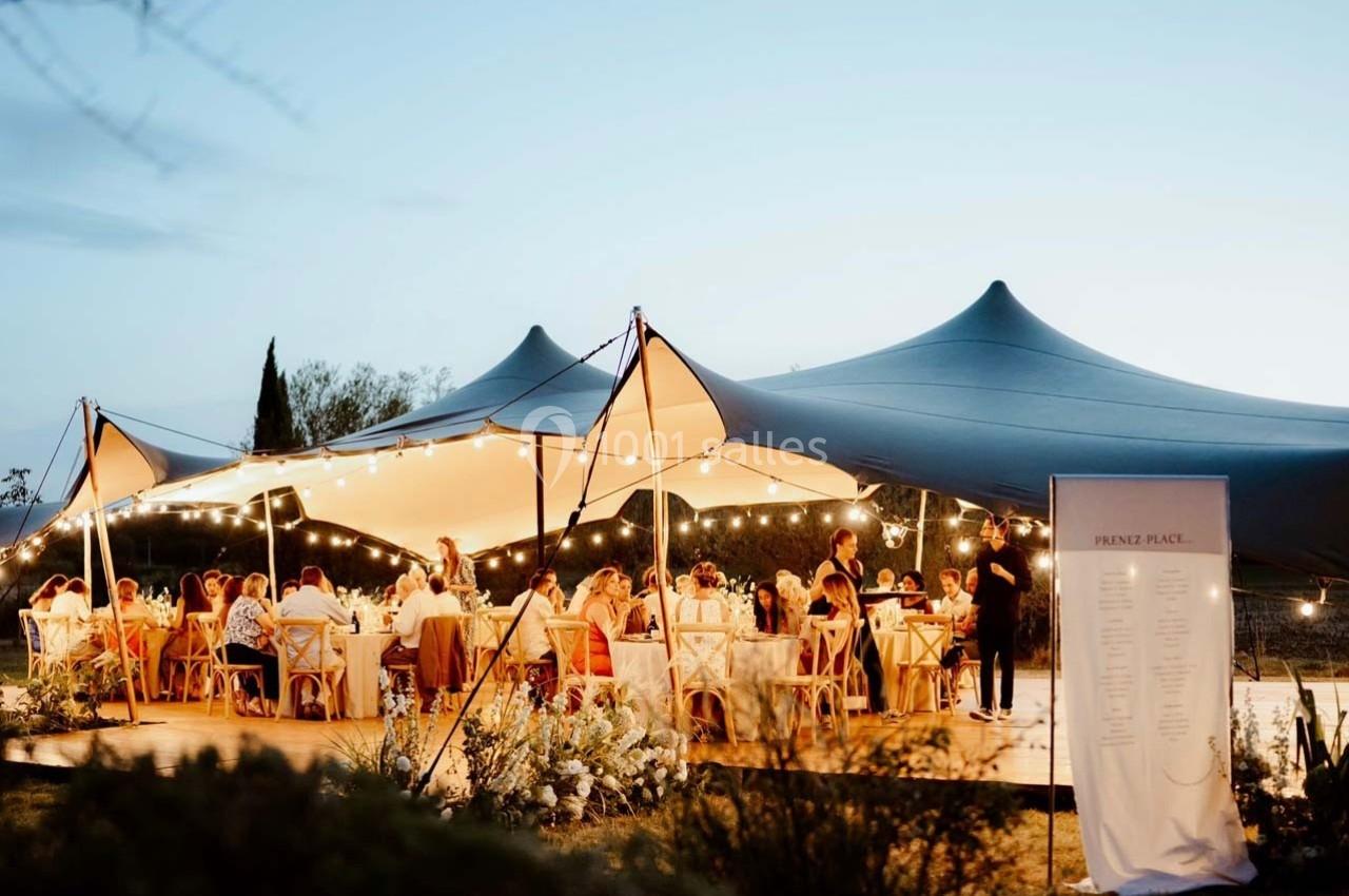 Dîner en plein air sous une tente éclairée par des guirlandes lumineuses, avec des invités assis à des tables décorées.