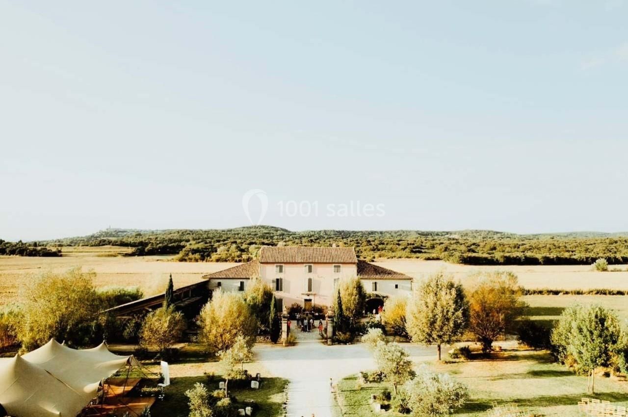 Vue aérienne d'une maison de campagne entourée de champs, d'arbres et de tentes dans un paysage rural ensoleillé.