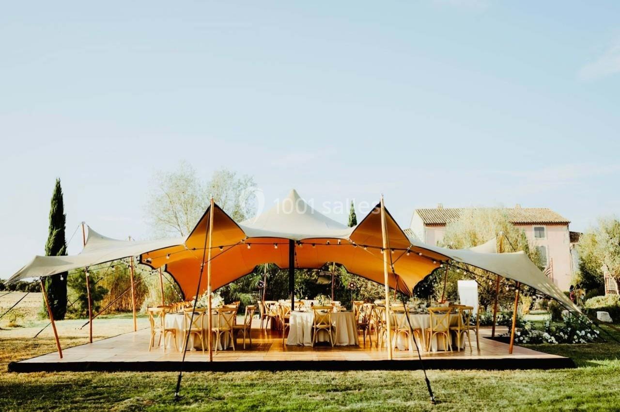 Tente beige installée sur une plateforme en bois, entourée de chaises et tables, dans un jardin ensoleillé.
