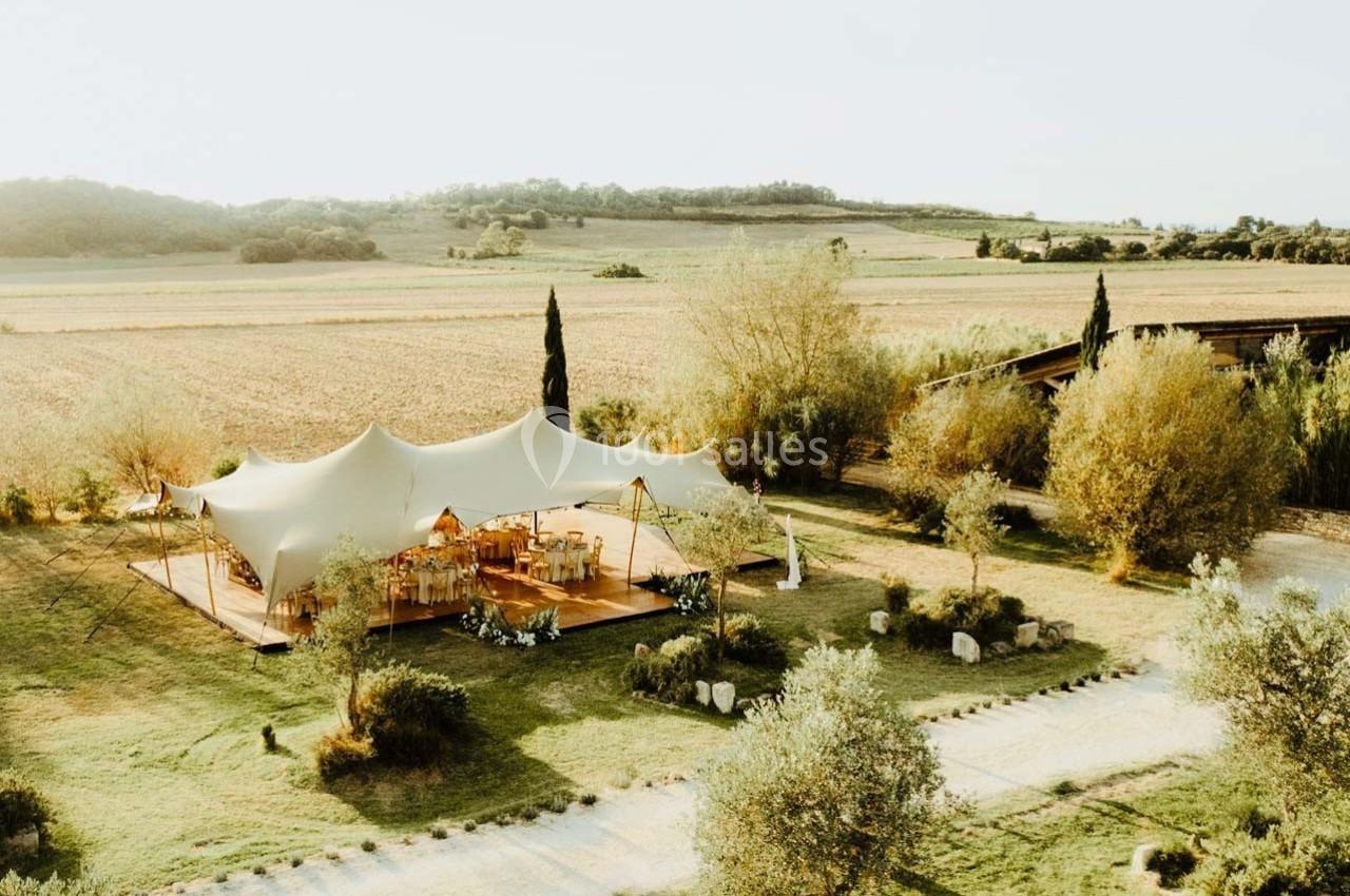 Tente blanche installée dans un champ entouré d'arbres, avec des tables et chaises disposées pour un événement.
