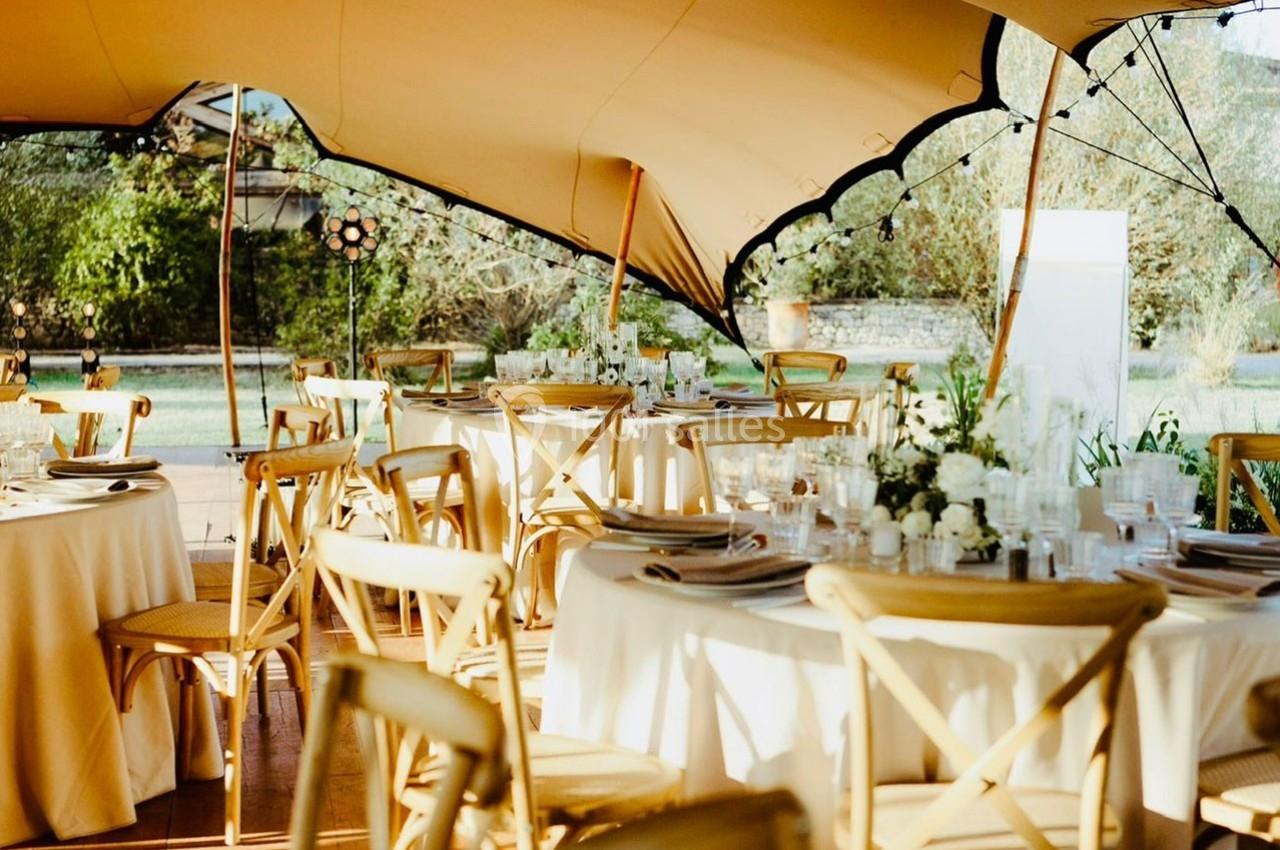 Tables rondes décorées avec vaisselle et fleurs sous une tente beige, entourées de chaises en bois clair.