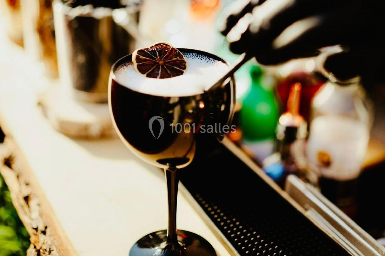 Un cocktail mousseux dans une coupe noire, décoré d'une tranche d'agrume séché, avec une main en arrière-plan.