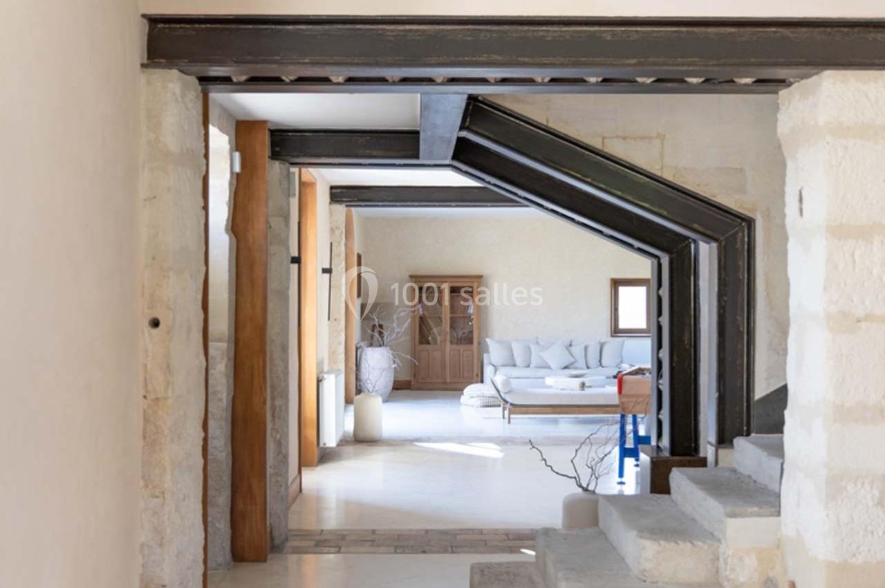 Vue intérieure d'une maison avec poutres métalliques apparentes, murs en pierre, et salon lumineux au fond.