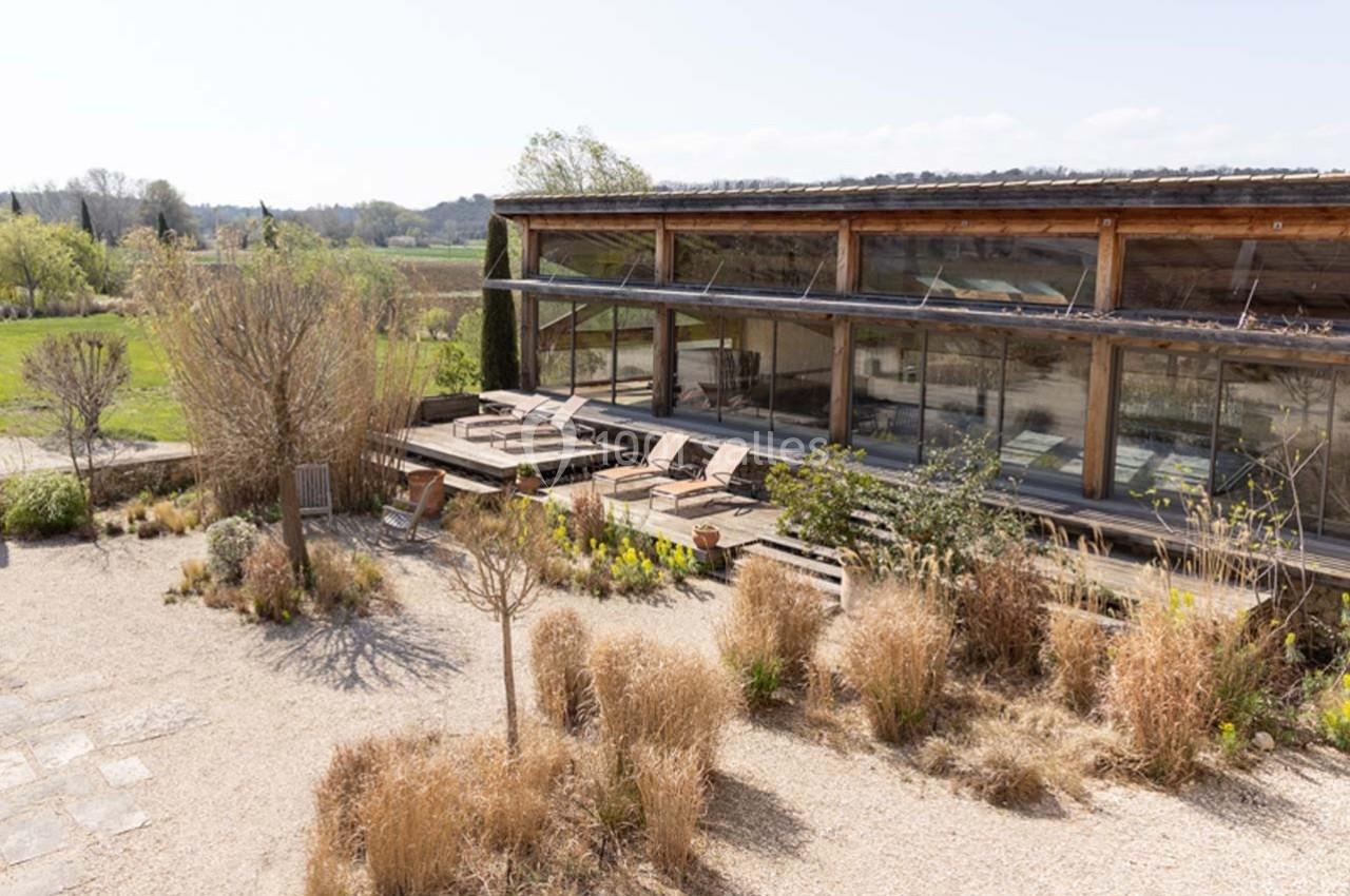 Bâtiment vitré entouré de végétation sèche, avec des chaises longues disposées sur une terrasse ensoleillée.