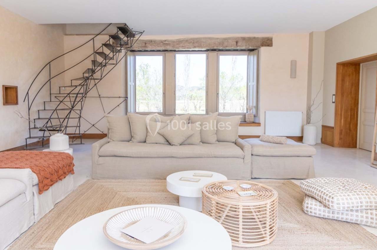 Salon lumineux avec canapé beige, tables basses blanches, escalier en métal et grandes fenêtres donnant sur l'extérieur.