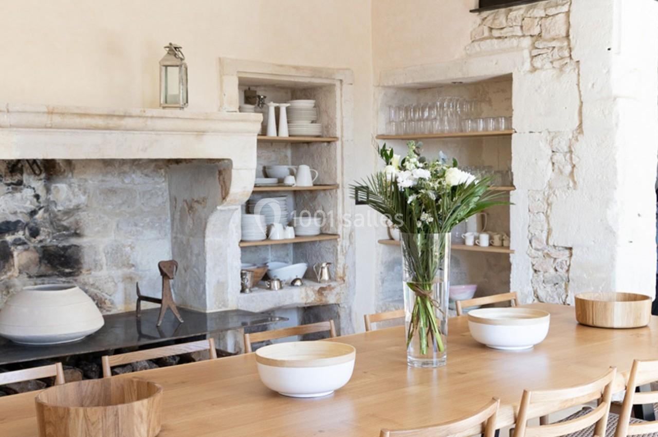 Grande table en bois entourée de chaises, décorée de fleurs, dans une cuisine rustique avec étagères et cheminée en pierre.