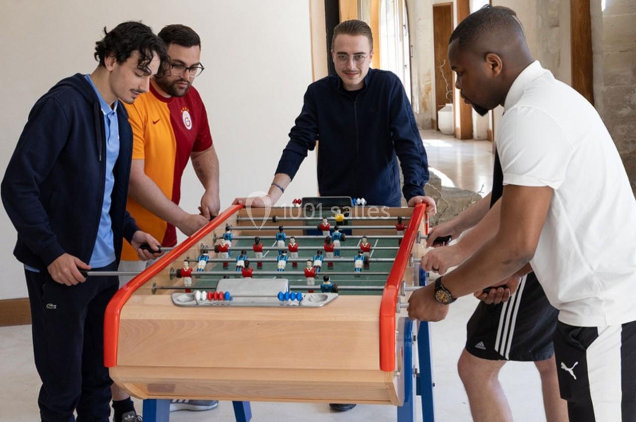 Quatre hommes jouent au baby-foot dans une pièce lumineuse avec des murs en pierre.