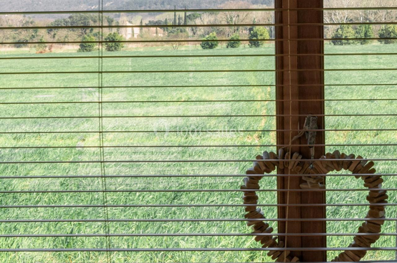 Vue d'un champ verdoyant à travers une fenêtre avec des stores en bois et une décoration en forme de cœur suspendue.