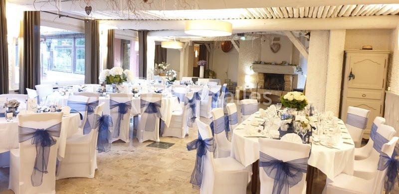 Salle de réception décorée pour un mariage, avec des tables rondes, nappes blanches et chaises ornées de nœuds bleus.