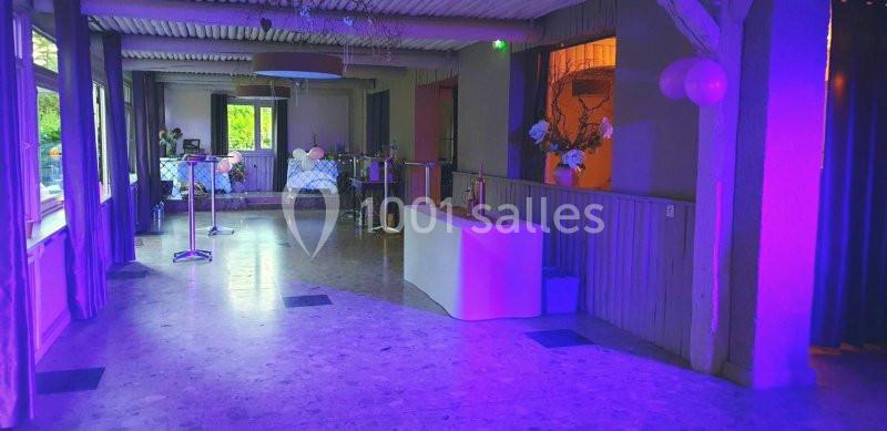 Salle de réception vide avec éclairage violet, tables hautes, décorations florales et ballons en arrière-plan.