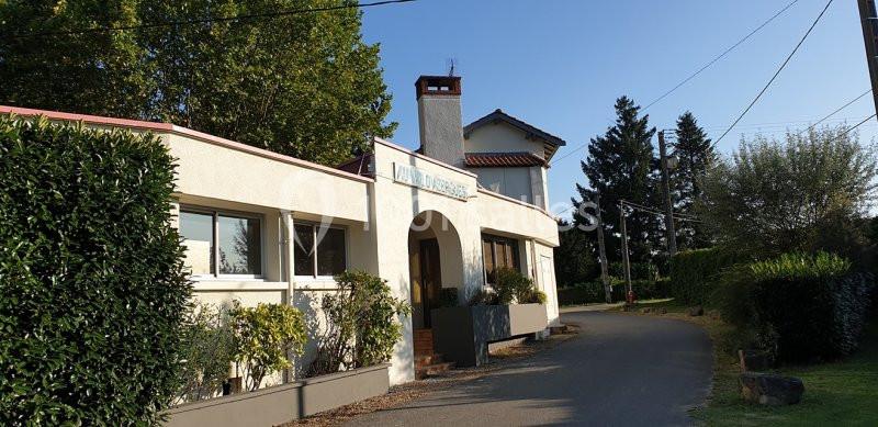 Façade d'un bâtiment blanc avec une arche, entouré de végétation, situé le long d'une petite route.