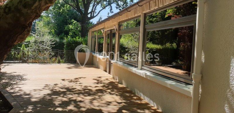 Terrasse en bois ombragée bordée de grandes baies vitrées donnant sur un jardin verdoyant.