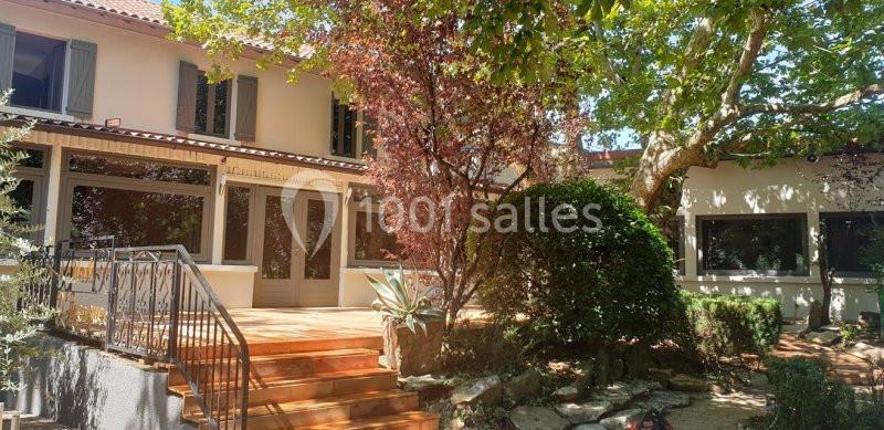 Maison avec terrasse en bois, entourée de végétation et d'arbres, sous une lumière ensoleillée.