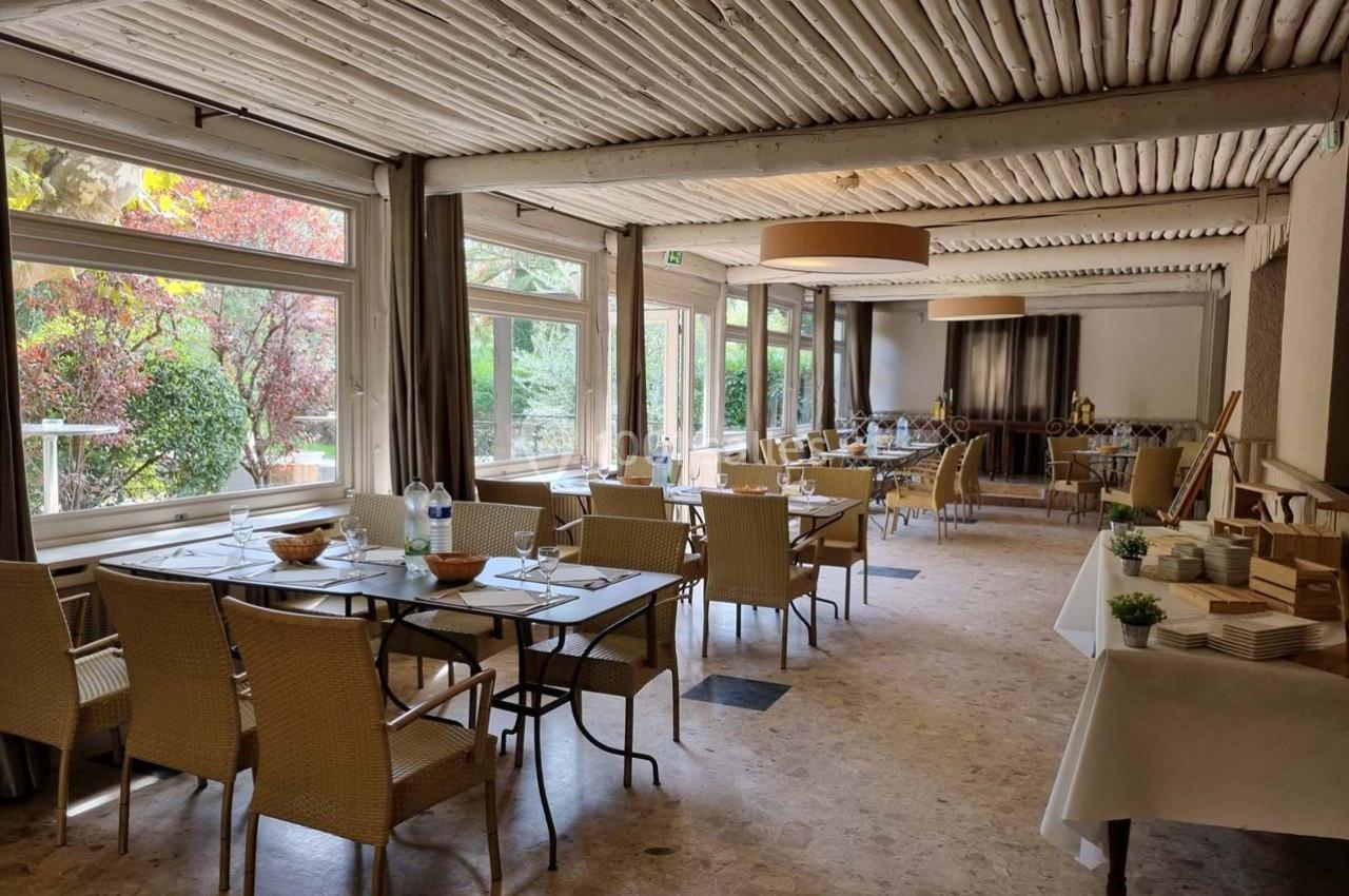 Salle lumineuse avec grandes baies vitrées, tables dressées et buffet disposé sur le côté, vue sur un jardin verdoyant.