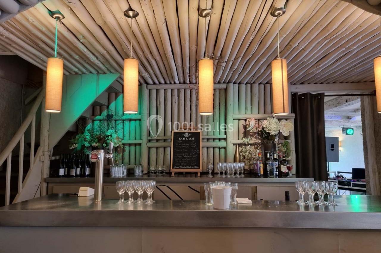 Comptoir de bar avec verres alignés, tableau noir au centre, décor en bois et suspensions lumineuses.