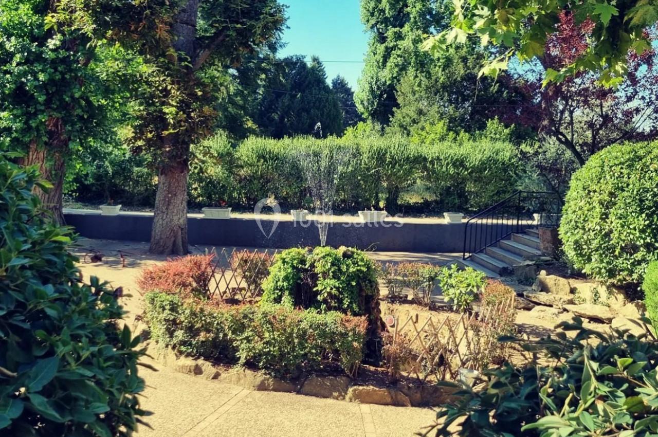 Jardin arboré avec une fontaine centrale entourée de buissons, allées pavées et escaliers en arrière-plan.