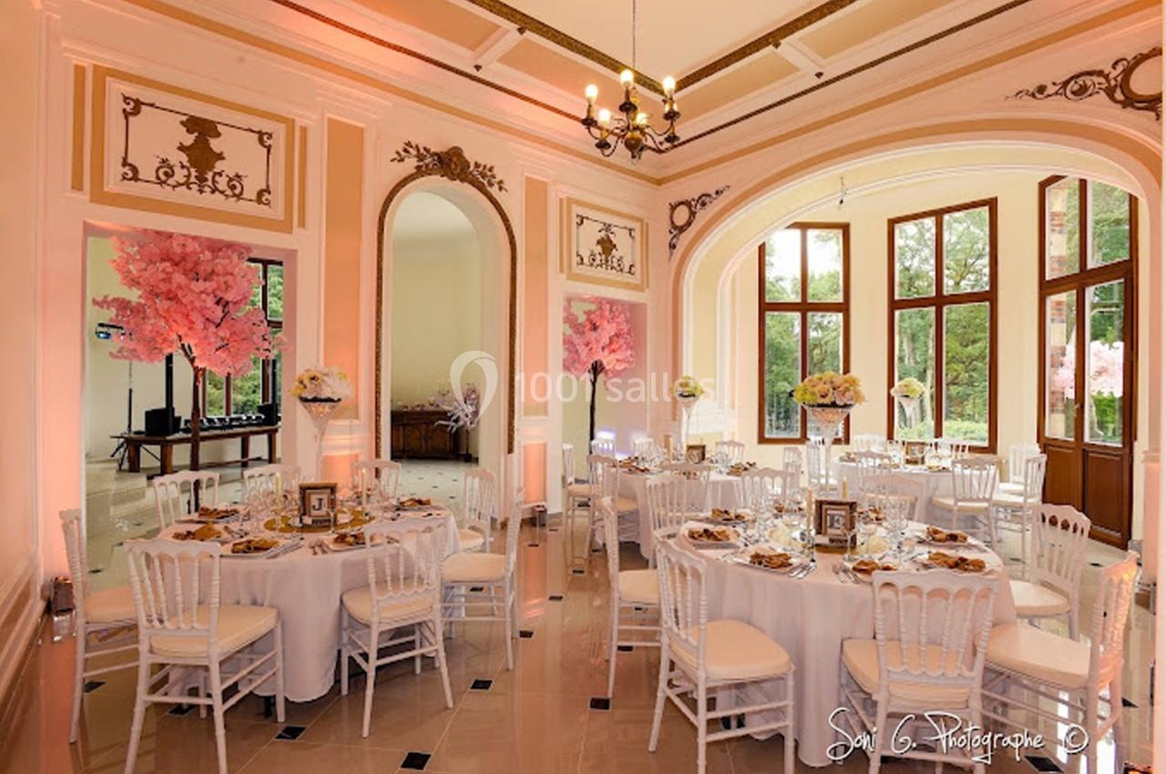 Salle de réception élégante avec tables rondes dressées, chaises blanches et décorations florales roses.