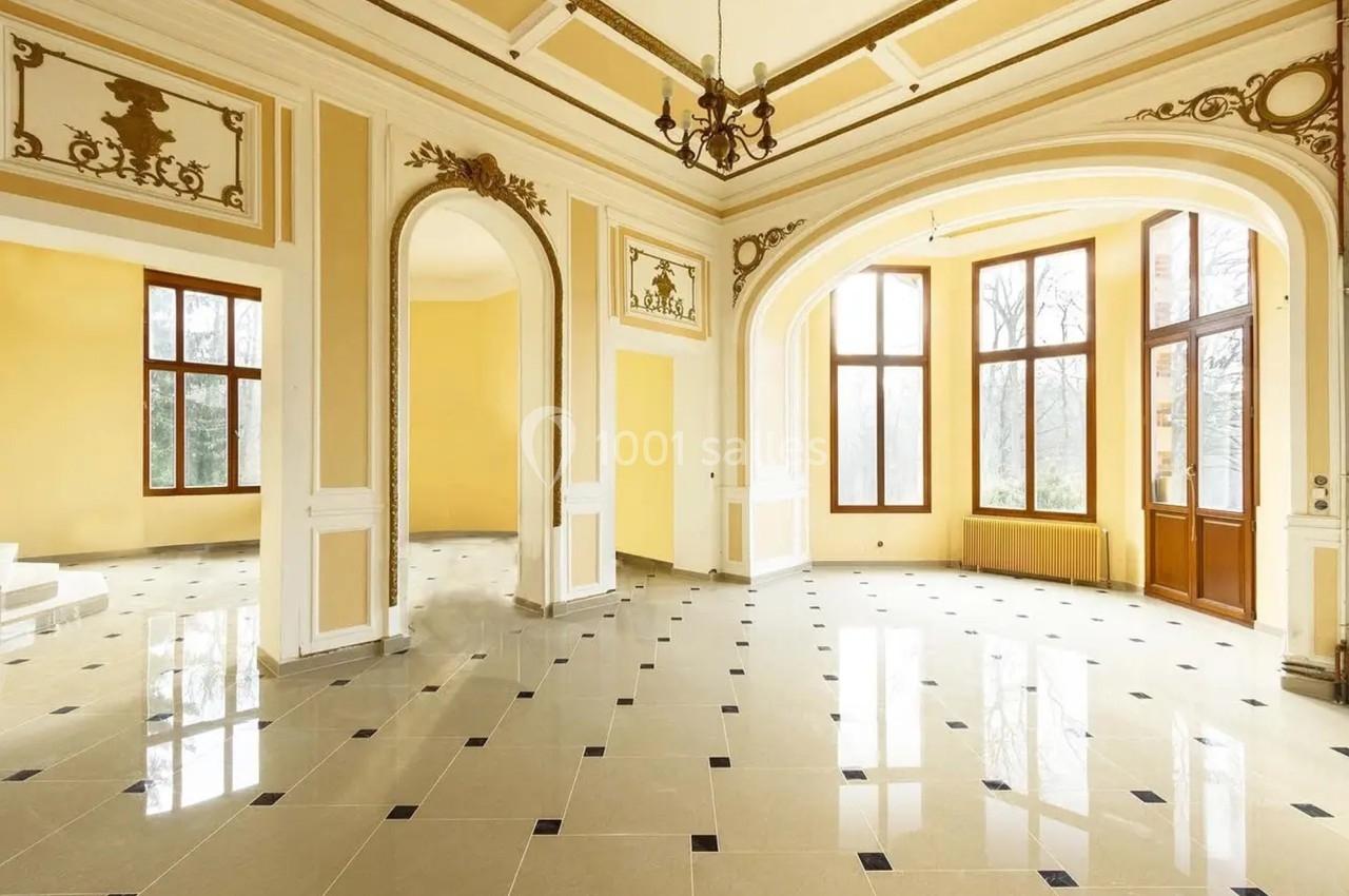 Salle lumineuse avec grandes fenêtres, carrelage beige et noir, moulures décoratives et hauts plafonds.