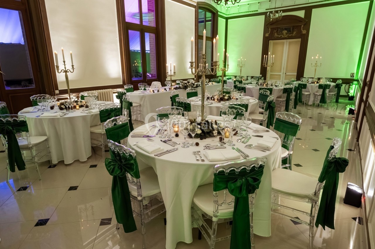 Salle de réception élégante avec tables rondes dressées, chaises ornées de nœuds verts et éclairage d'ambiance.