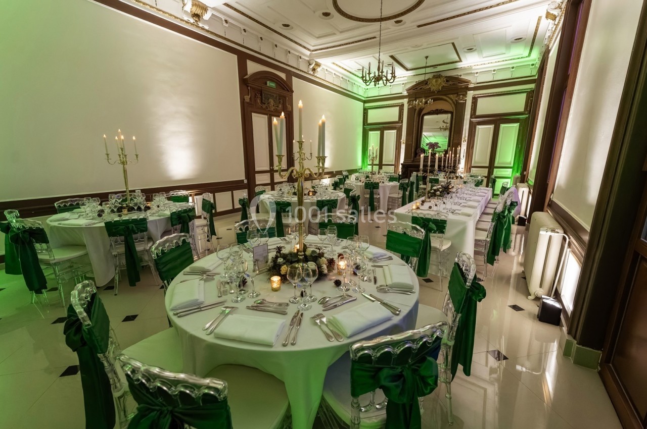 Salle de réception élégante avec tables rondes dressées, nappes blanches, chaises décorées de nœuds verts et chandeliers.